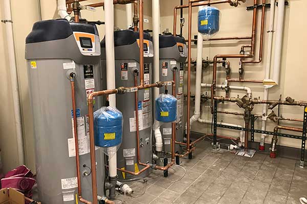 Row of tall water heaters with blue expansion tanks and copper piping in a utility room